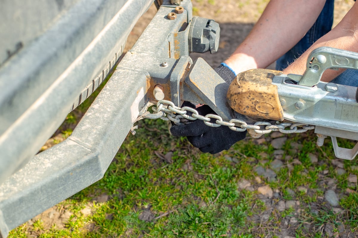 A man attaches a trailer to a car, securing safety chains to the trailer's hitch and drawbar